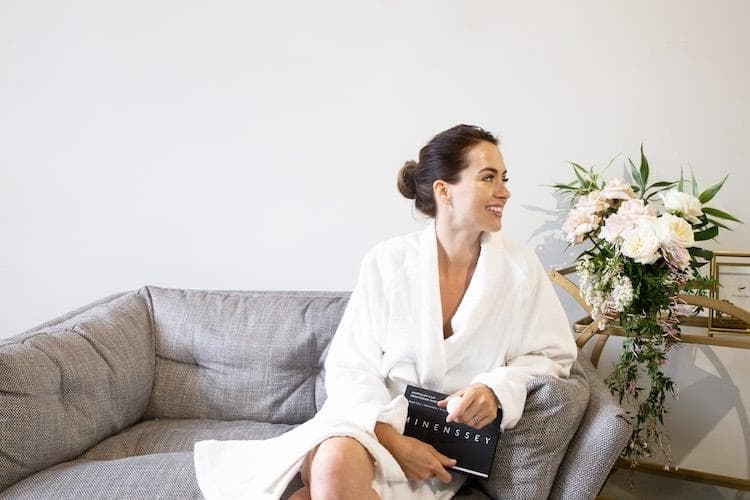 Woman with clay face mask applied, resting on massage bed with flowers and skincare products nearby