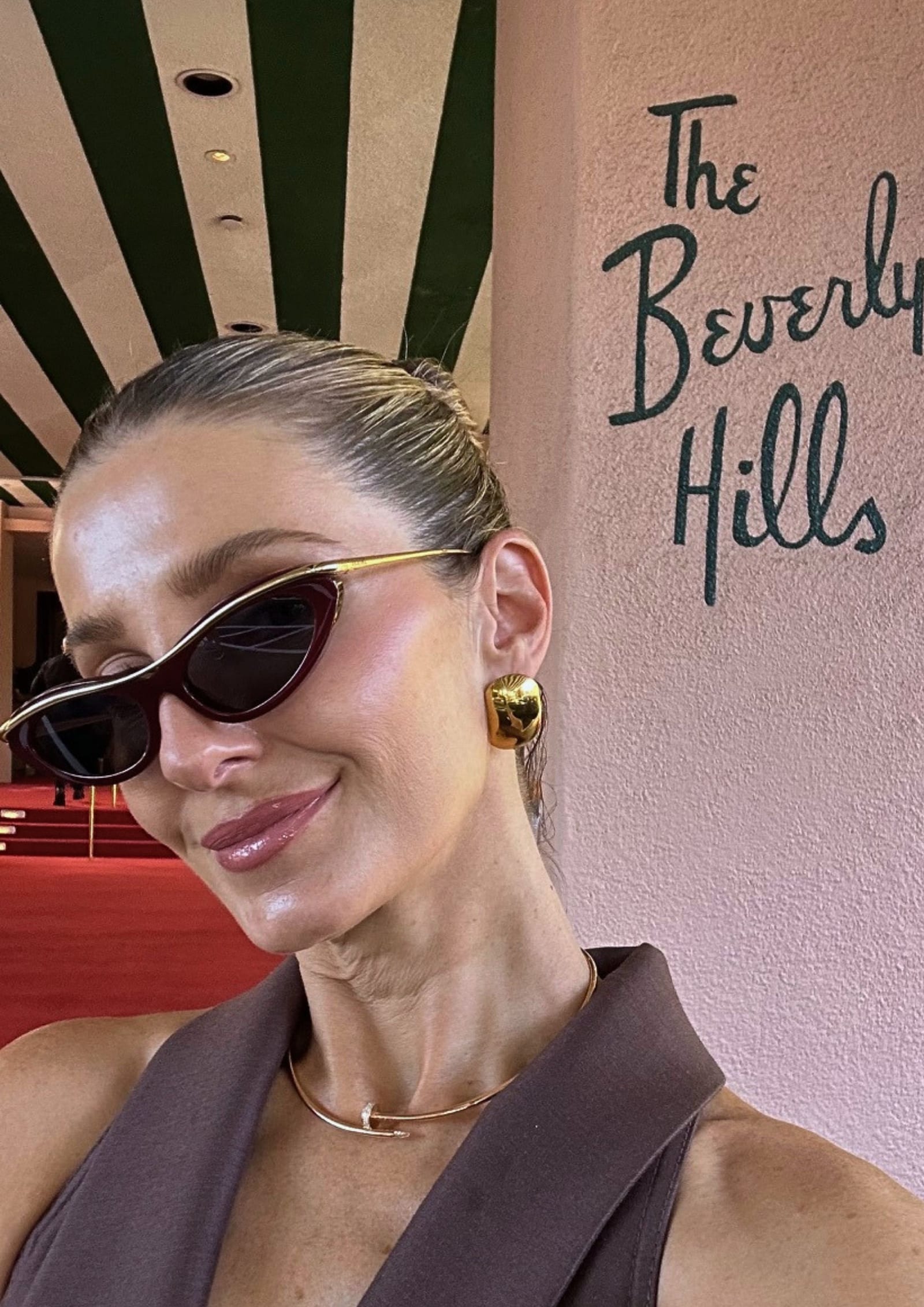 Kate Waterhouse taking a selfie outside The Beverly Hills Hotel beside the pink wall with green script lettering, wearing tortoiseshell cat-eye sunglasses and gold statement earrings