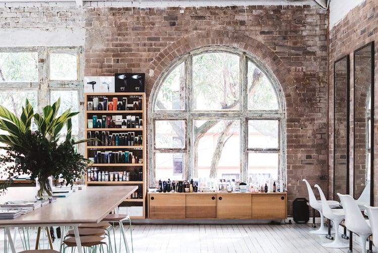 Bright, minimalist hair salon interior with vaulted ceilings, white flooring, natural light and communal wooden table