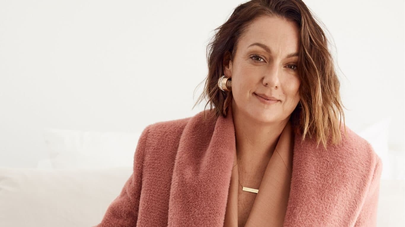 Celeste Barber smiling in a blush pink coat with gold jewellery, photographed for Beautiful Inside by Beauticate