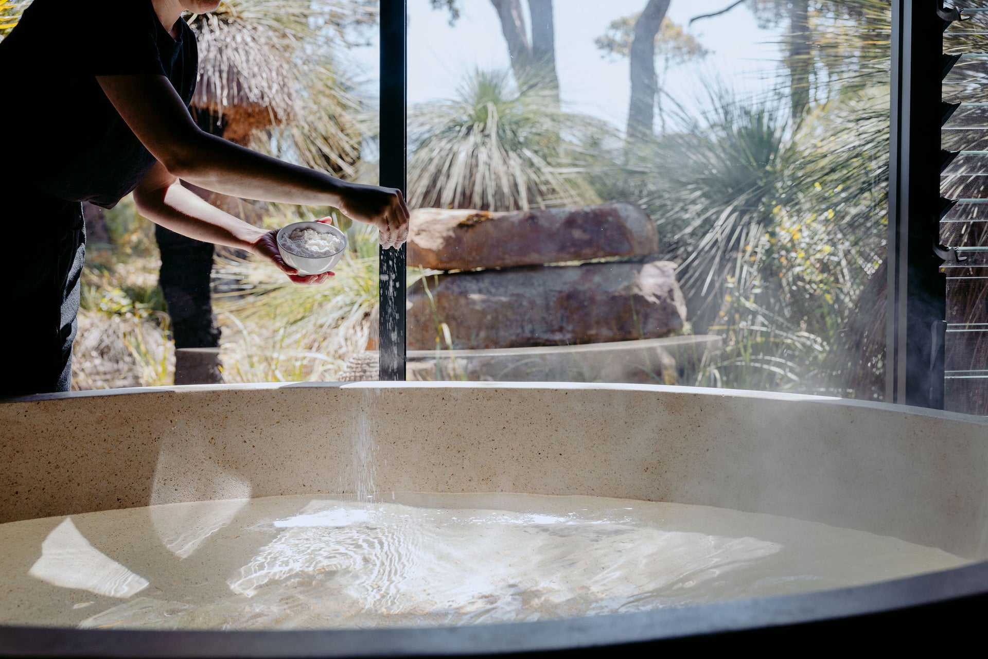 A therapist sprinkles bath salts into a stone soaking tub overlooking native bushland at Yala Yallingup's spa