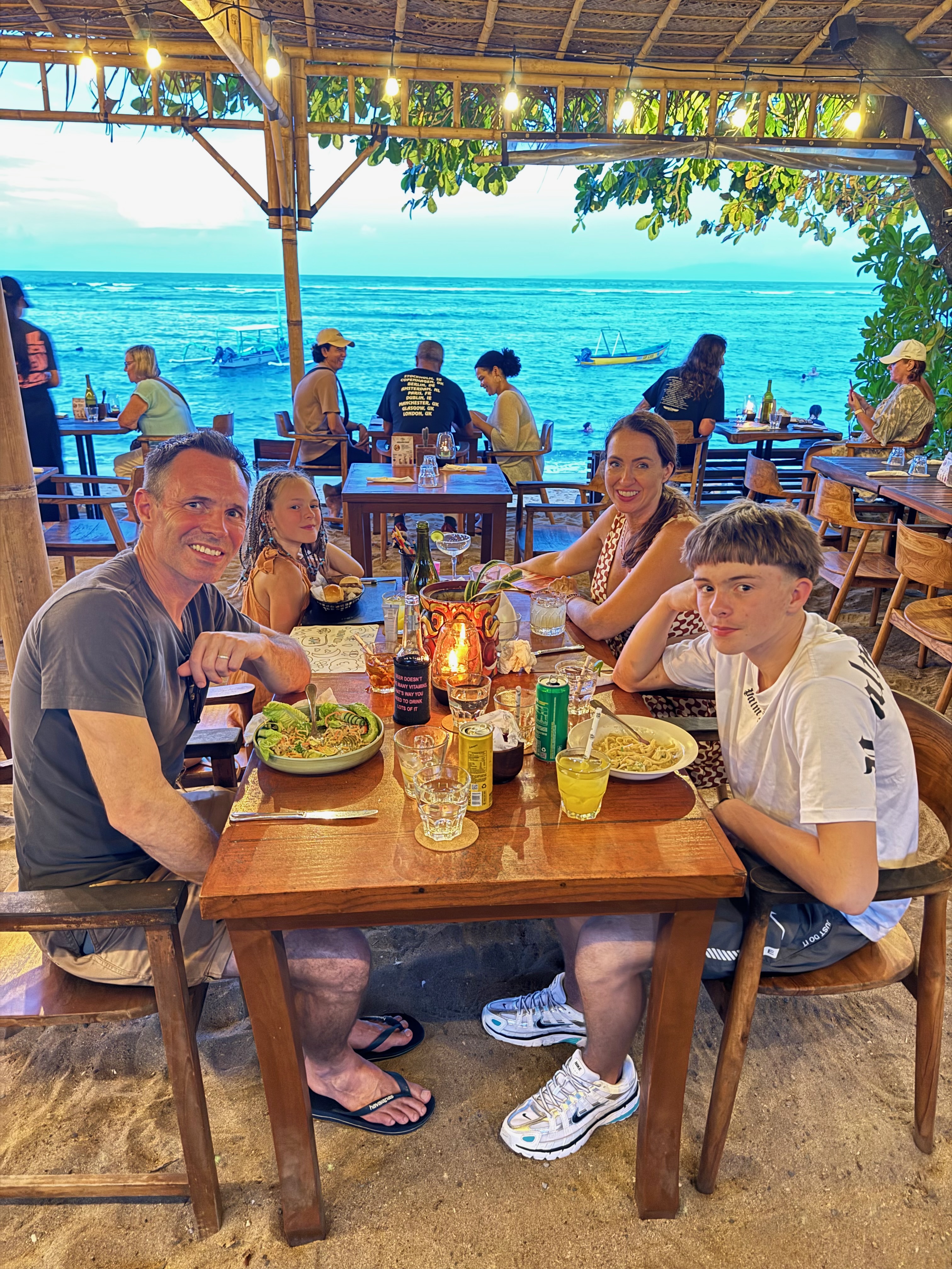 A beachfront dinner table set on the sand in Sanur at dusk, with warm lighting and the ocean behind