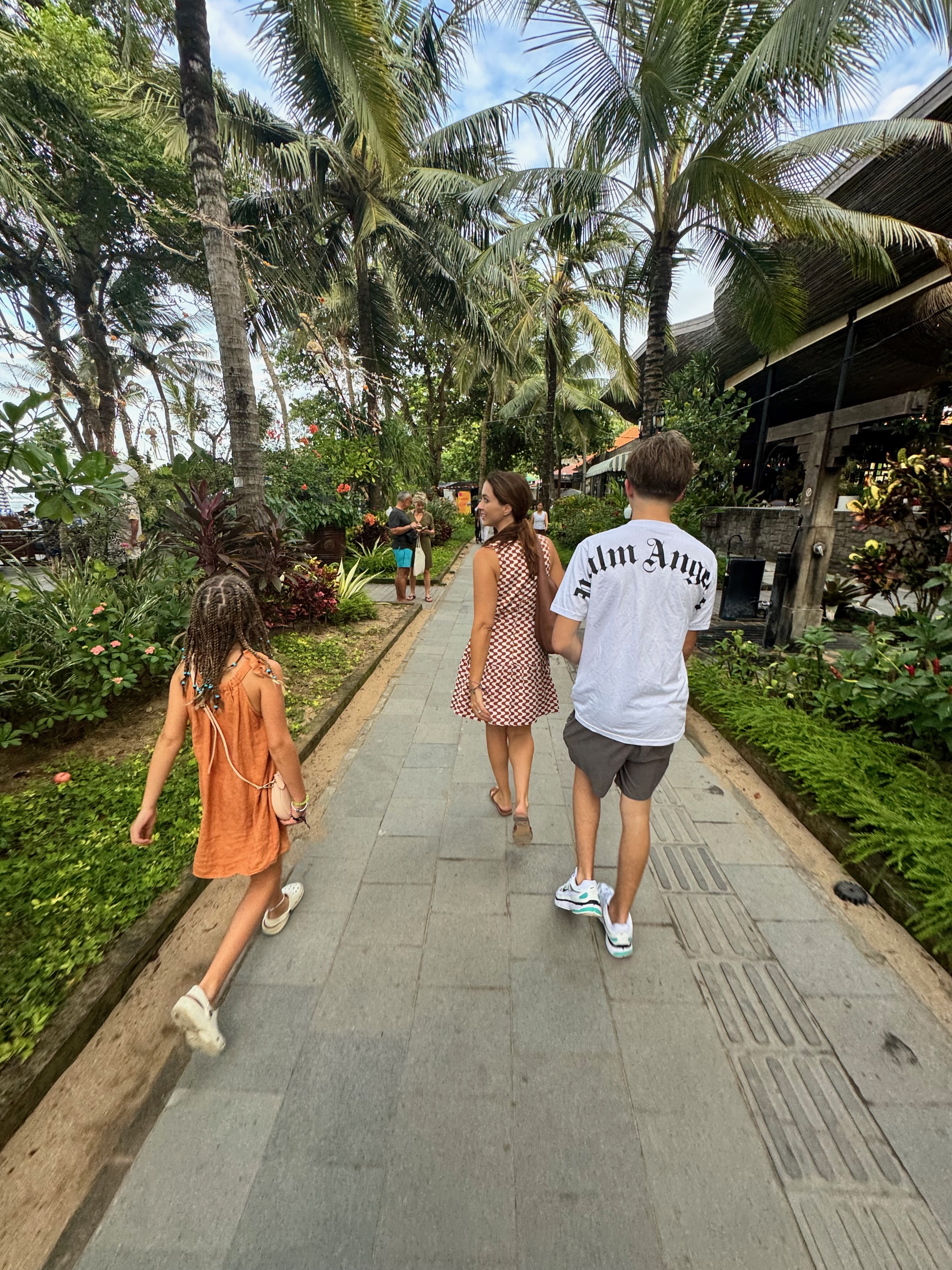 A family walking along the wide, flat Sanur beach path lined with tropical trees and local warungs