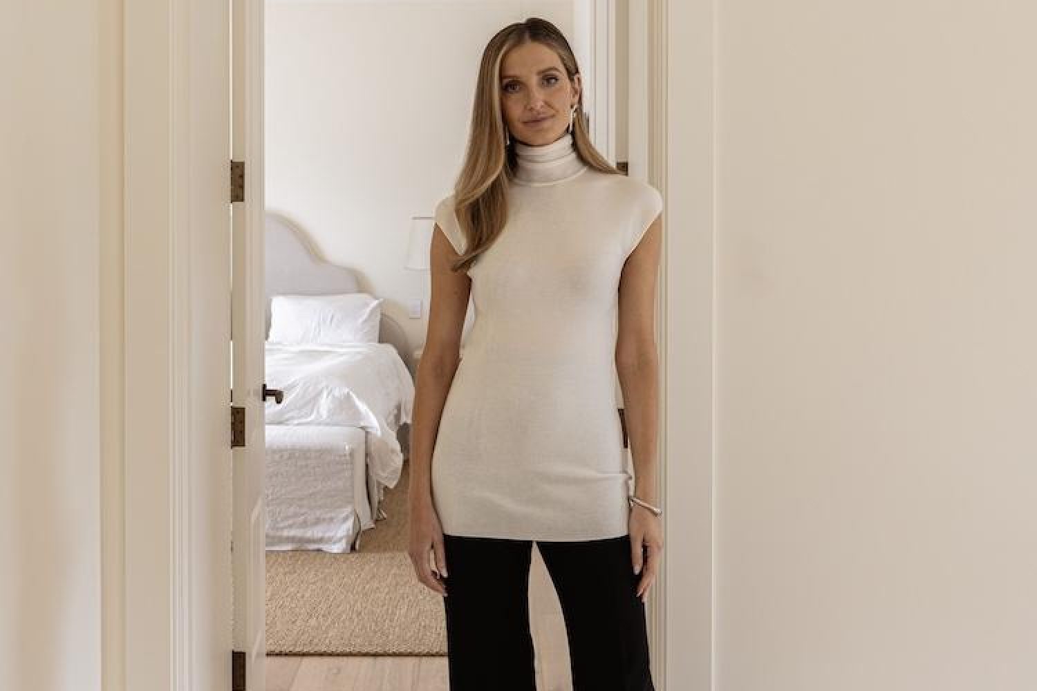 Kate Waterhouse in a cream sleeveless turtleneck and black trousers standing in a bedroom doorway