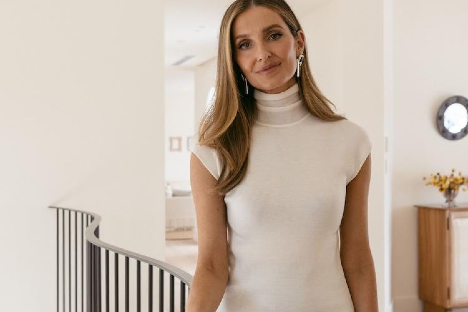 Kate Waterhouse in a cream cap-sleeve turtleneck top, smiling by a curved staircase with silver earrings