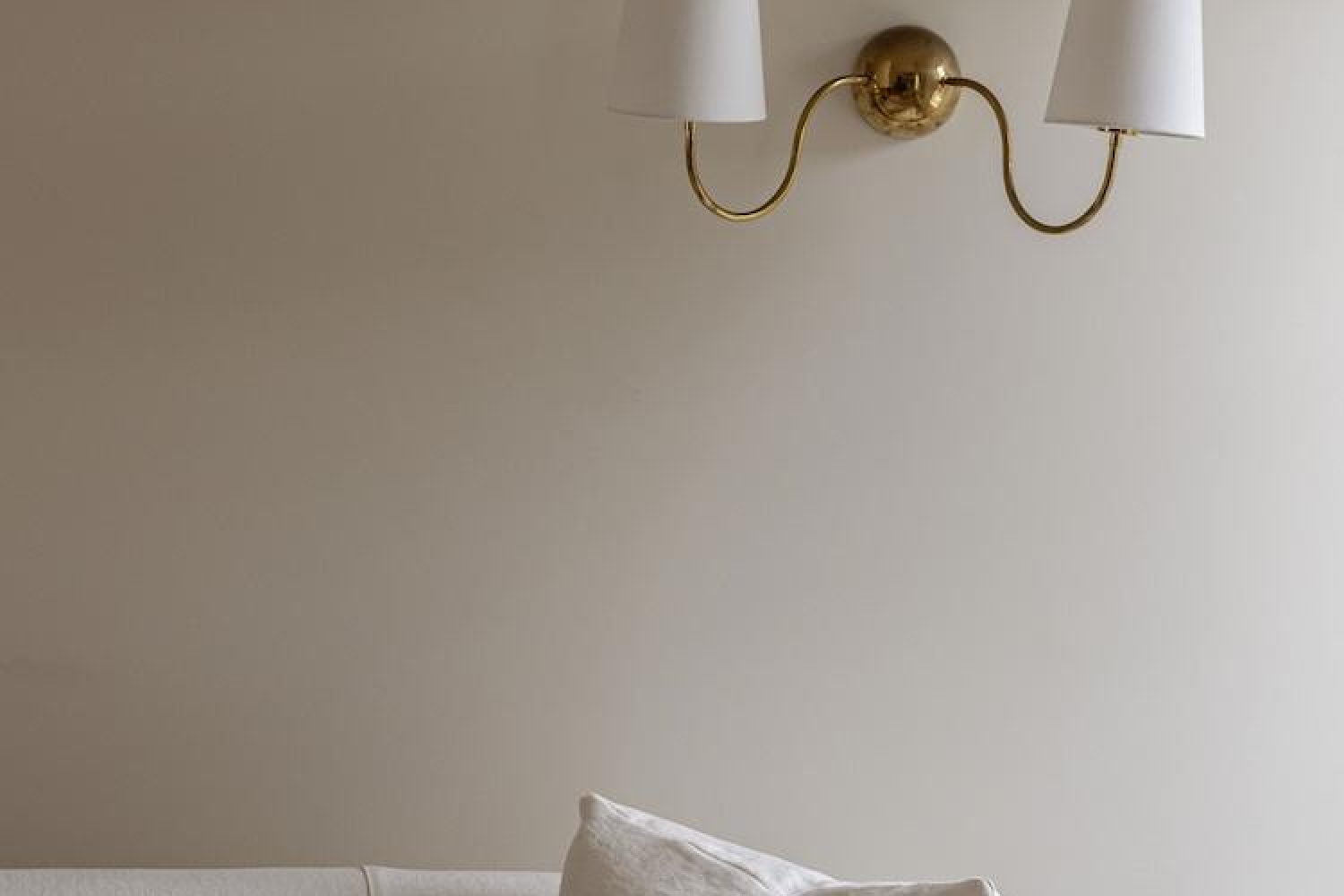 Brass double-arm wall sconce with white shades above a linen sofa cushion in a neutral-toned living room