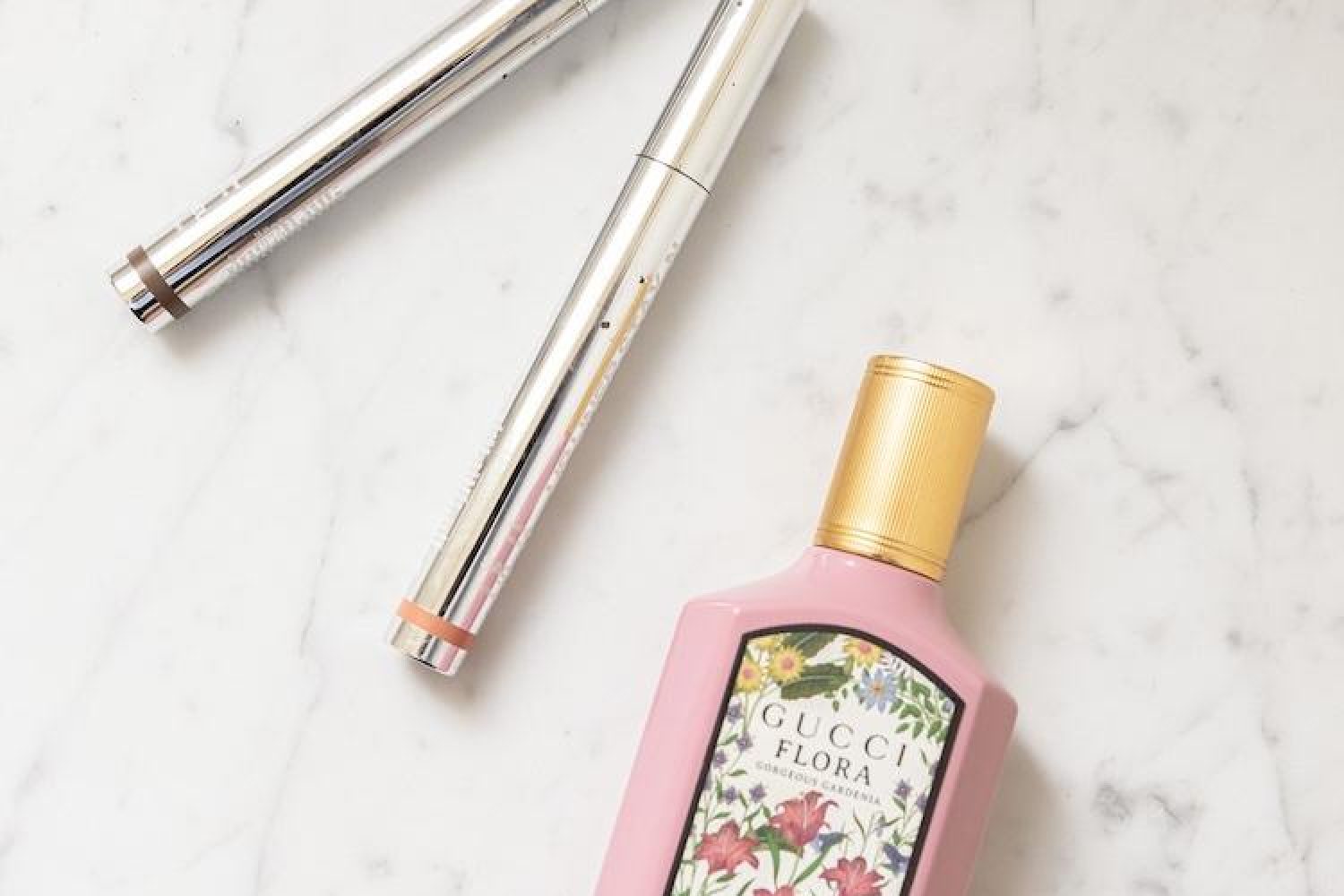 Gucci Flora Gorgeous Gardenia perfume in a pink bottle beside two silver YSL Touche Eclat pens on marble