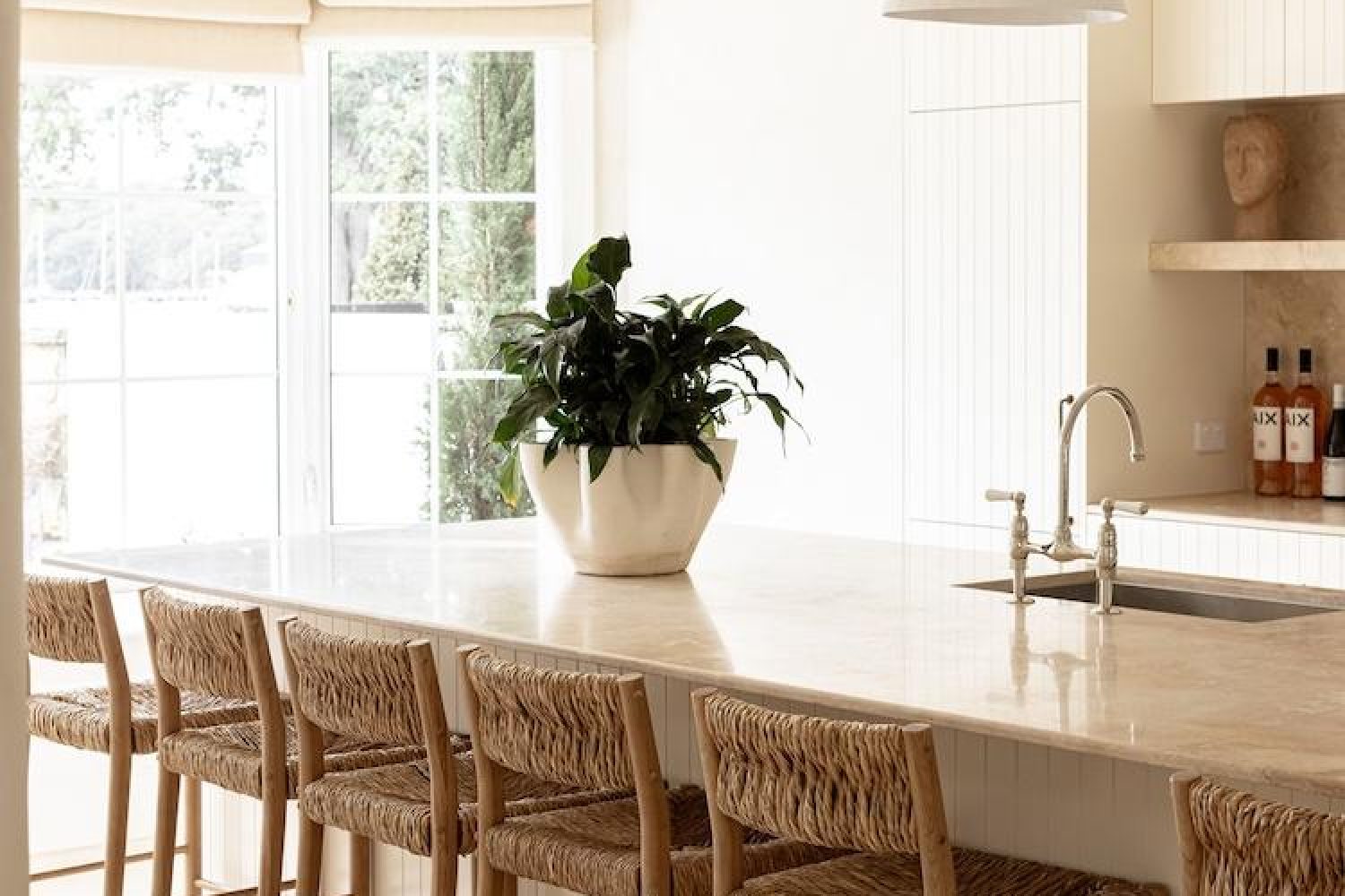 Light-filled kitchen island with woven rattan bar stools, a potted plant and bottles of AIX rose on the shelf