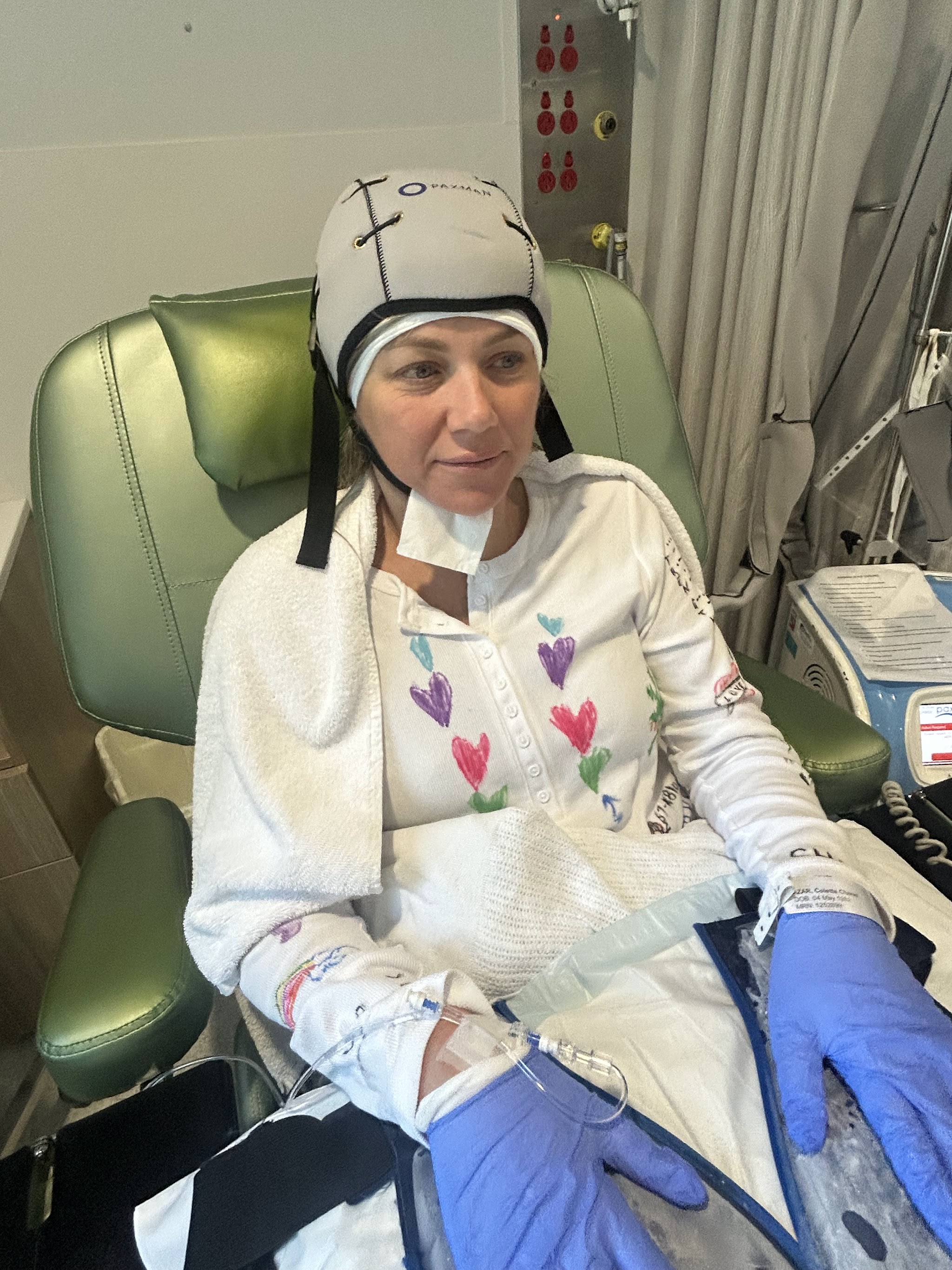 Colette during a chemotherapy session wearing the cold cap and a heart-print cardigan, IV line in her arm